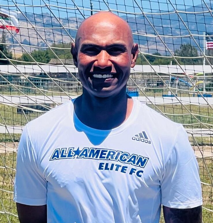 A bald man with a shaved head is standing in front of a soccer goal.